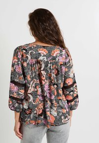 Blouse florale avec des motifs colorés sur fond noir, dotée de manches bouffantes et d'un dos plissé. Tissu léger et texturé.