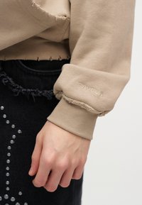 Manche de sweat-shirt beige avec bords bruts et "FAVELA" brodé sur le poignet, portée sur un jean noir déchiré orné de clous métalliques.