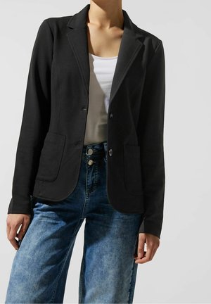 Femme portant un blazer noir boutonné, un haut blanc et un jean bleu taille haute, debout devant un fond clair uni.