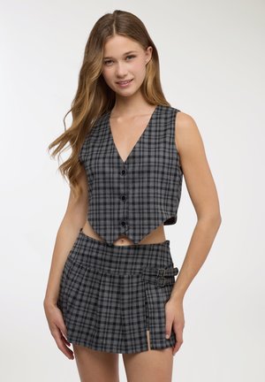 Gilet corto a quadri grigi con scollo a V profondo e chiusure con bottoni, abbinato a una minigonna plissettata coordinata con fibbie laterali.