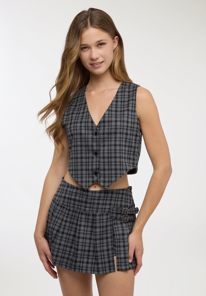 Gilet corto a quadri grigi con scollo a V profondo e chiusure con bottoni, abbinato a una minigonna plissettata coordinata con fibbie laterali.