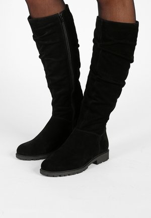 Stivali neri in suede al ginocchio con cerniera laterale indossati sopra collant neri trasparenti su gambe in piedi su uno sfondo bianco.