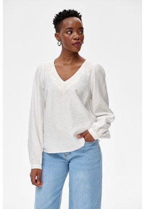 Kobieta o krótkich włosach, ubrana w białą bluzkę z długim rękawem i jasnoniebieskie jeansy, patrząca w prawo, z jedną ręką w kieszeni.