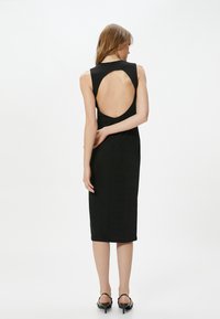 Schwarzes ärmelloses Kleid mit hohem Kragen und großem kreisförmigen Ausschnitt im Rücken. Figurbetonte Silhouette, aus glattem Stoff, knielang.
