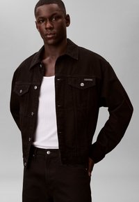 Hombre con chaqueta de mezclilla negra sobre camiseta sin mangas blanca de canalé, con pantalones negros, de pie frente a un fondo gris liso.