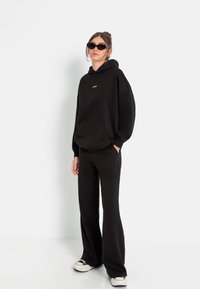 Felpa nera con tasca frontale e logo LSCW; abbinata a pantaloni neri larghi. Il modello indossa occhiali da sole neri e sneakers con accenti bianchi.