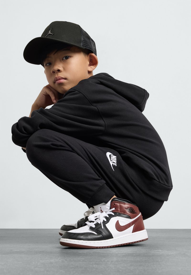 Črna kapuca s kapuco in trenirka z belim Nike logotipom, črne in rdeče teniske, črna bejzbolska kapa, v poklonskem položaju na sivi talni oblogi.