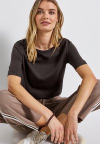 Černý top s krátkým rukávem a kulatým výstřihem, nošený s béžovými tepláky se bočními pruhy. Model sedí, viditelná světlá textilie.