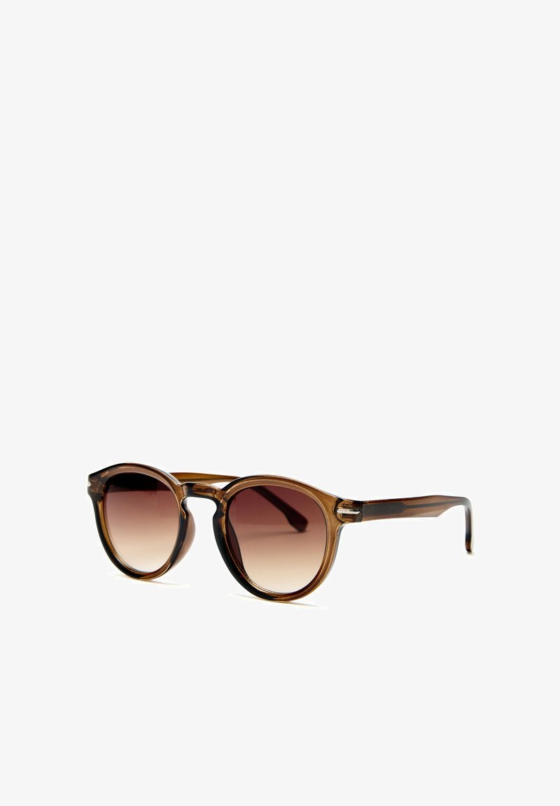 Lunettes de soleil rondes transparentes marron avec verres dégradés foncés posées sur une surface blanche, inclinées vers la droite.