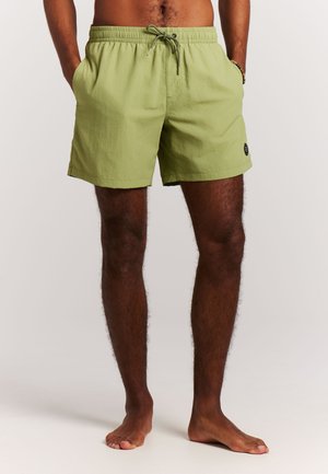 Man draagt lichtgroene zwembroek met trekkoord, staat blootsvoets met handen in de zakken tegen een effen achtergrond.