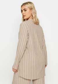Maglietta beige a maniche lunghe con righe verticali blu navy, tessuto testurizzato, spacco laterale e collo alto. Indossata con un paio di pantaloni abbinati.