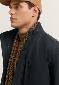 Dunkle, strukturierte Jacke mit hohem Kragen und Reißverschlusstasche, über einem braunen gemusterten Schal und einem braunen Rollkragenpullover getragen, kombiniert mit einer Mütze.