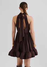 Brown halter dress with a tied neckline, open back, and a flared skirt featuring ruffled hem. Made of a smooth, textured fabric.
