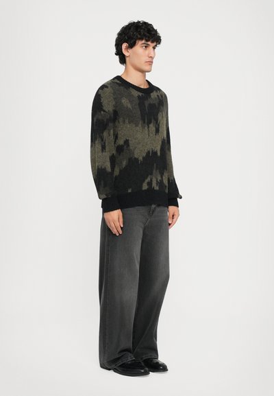 Gebreide trui in groen en zwart, met een gebroken patroon. In combinatie met losse donkergrijze jeans en zwarte schoenen.