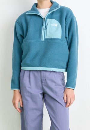 Persoon die een teal fleece trui draagt met een ritssluiting op de borstzak en lichtpaarse broek, staand tegen een effen witte achtergrond.