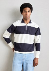 Pullover a righe blu navy e bianche con colletto con zip e polsini a coste. Realizzato in tessuto morbido, presenta un logo ricamato sul petto.