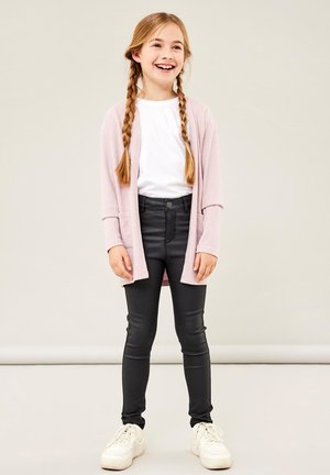 Cardigan rosa chiaro con una vestibilità ampia, t-shirt bianca, pantaloni neri aderenti e sneakers bianche, mostrano trecce e uno sfondo neutro.