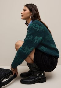 Zeleni pleteni pulover z teksturiranim vzorcem, nošen čez črno krilo. Črni gležnarji s srebrnimi dodatki, kombinirani s tkanim torbico.