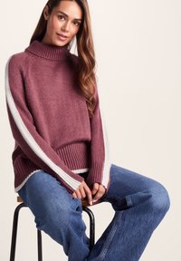 Maroon knitted sweater with a high collar and white striped sleeves, featuring a ribbed hem. Paired with blue denim jeans.