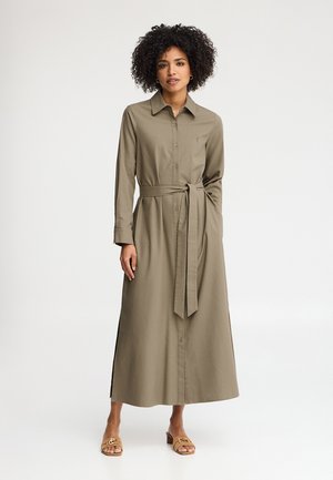 Kobieta o kręconych włosach, ubrana w khaki sukienkę koszulową na długi rękaw, z guzikami i paskiem w talii oraz beżowe sandały na obcasie.