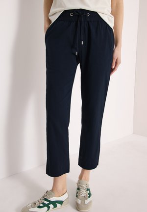 Personne portant un pantalon à cordon bleu marine et des baskets blanches avec des rayures vertes, debout devant un fond clair uni.