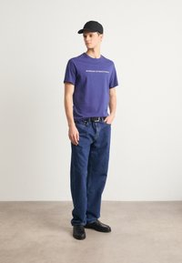 Purple cotton t-shirt with "BARBOUR INTERNATIONAL" printed in white, paired with loose-fitting blue denim jeans and black shoes.