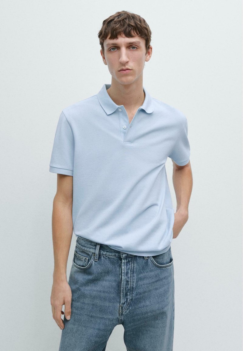 Junger Mann mit kurzem braunem Haar, der ein hellblaues Poloshirt und blaue Jeans trägt und vor einem schlichten weißen Hintergrund steht.