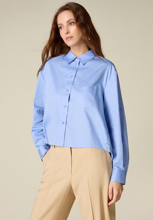 Kvinde iført en løs lyseblå skjorte med knapper og forreste lomme samt beige højttaljede bukser, stående med hænder i lommerne mod en ensfarvet baggrund.