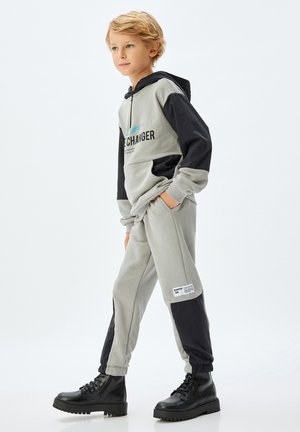 Blond chłopiec chodzi, nosząc szarą i czarną bluzę z kapturem oraz pasujące joggery z czarnymi butami wojskowymi na białym tle.