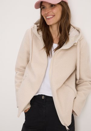 Light beige fleece jacket with a hood, front zipper closure, and side pockets. Worn over a white shirt and black pants, featuring textured fabric.