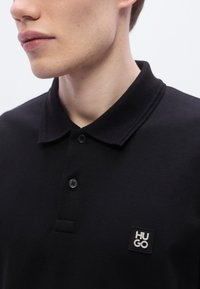 Μαύρο πόλο πουκάμισο από υφασμάτινη υφή που διαθέτει γιακά, τρία κουμπιά στο ντεκολτέ και ετικέτα με την επωνυμία "HUGO" στο αριστερό στήθος.