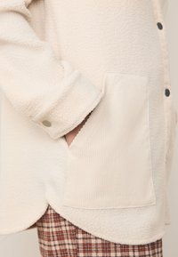 Camicia in pile color crema con orlo arrotondato, chiusura frontale con bottoni e tasca in velluto a coste a contrasto. Tessuto strutturato e maniche arrotolate. Pantaloni a quadri visibili sotto.