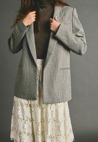 Blazer gris à chevrons avec épaules structurées, associé à une jupe en dentelle crème ornée de motifs floraux et de bords festonnés.