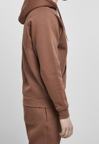 Hnědá mikina s kapucí a hladkým povrchem, vybavená klokaní kapsou a žebrovanými manžetami. Stylizováno s odpovídajícími kalhotami pro jednotný vzhled.