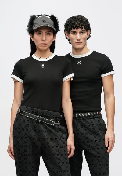 Juodos prigludusios marškinėliai su baltais apvadais ir mažyliu logotipu. Tamsūs raštuoti kelnės su sidabriniais kuoleliais papuoštu diržu ir juoda kepure su kailio akcentu.