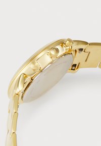Montre en acier inoxydable doré avec un boîtier incurvé, un bracelet lisse et deux boutons latéraux. L'arrière affiche le logo et les détails du matériau.