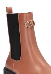 Stivale alla caviglia in pelle marrone con pannello elastico laterale nero, emblema dorato sulla parte anteriore superiore e linguetta sul retro.