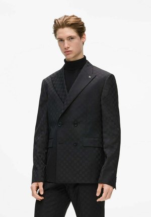 Homme portant un blazer croisé à carreaux noirs et un col roulé noir, debout devant un fond blanc uni.
