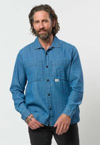 Homme aux cheveux ondulés et à la barbe, portant une chemise en jean bleu boutonnée et un pantalon noir, debout et souriant devant un fond uni clair.