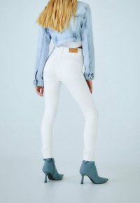 Jean skinny blanc taille haute avec ourlets effilochés aux chevilles, associé à une veste en denim bleu clair raccourcie et des bottines à talons hauts bleues.