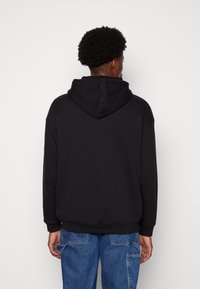 Felpa nera con cappuccio regolabile e polsini a coste. Il design presenta un tessuto morbido e testurizzato e una vestibilità comoda. Indossata con jeans cargo blu.