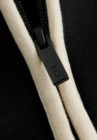 Sort stof med creme lynlåsdetalje. Lynlåsens trækstykke har en sort flap med et præget bogstav "B." Nærbillede fremhæver tekstur og design.