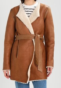 Abrigo de cuero marrón con cuello de piel vuelta y cinturón, con mangas largas y bolsillos laterales. El interior tiene una textura suave.