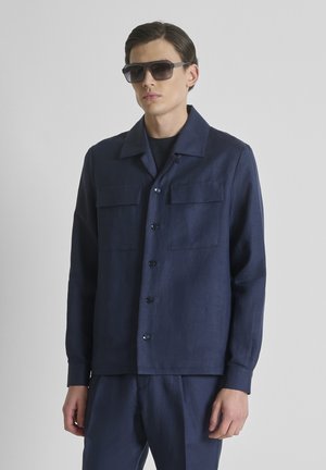 Homme portant une veste boutonnée bleu marine foncé et un pantalon assorti avec de grandes lunettes de soleil noires, debout sur un fond uni gris clair.