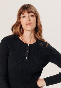 Zwarte geribbelde gebreide top met lange mouwen en een ronde halslijn, voorzien van een knoopsluiting met drie knopen aan de voorkant en aansluitende manchetten.
