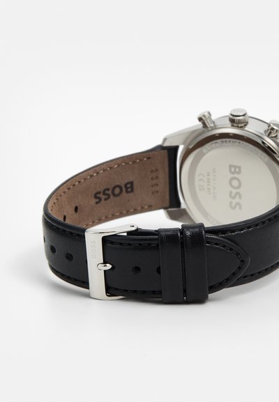 Bracelet de montre en cuir noir avec boucle en métal argenté, présentant un design cousu et un dessous de marque. Boîtier en acier inoxydable visible.
