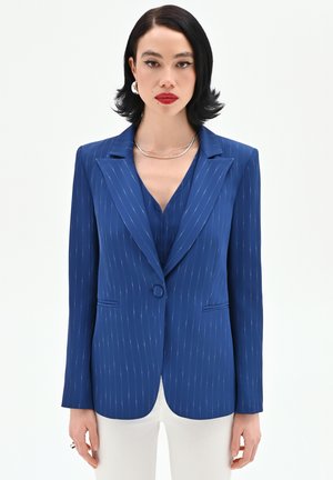 Donna con capelli corti neri che indossa un blazer a righe blu su un top abbinato e pantaloni bianchi, accessoriata con collana e orecchini d'argento.