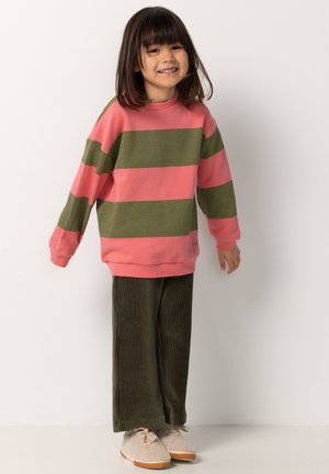 Un jeune enfant souriant, portant un pull rayé rose et vert, un pantalon large vert olive et des baskets beige en fausse fourrure, se tenant devant un mur blanc.
