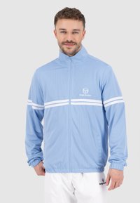 Giacca a rete blu chiaro con strisce orizzontali bianche, chiusura con zip davanti, colletto alto e polsini elastici. Presenta il logo sul lato sinistro del petto.