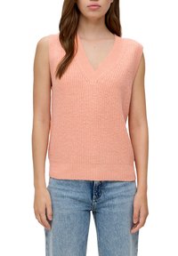 Pfirsichfarbener, gestrickter ärmelloser Pullover mit V-Ausschnitt und geripptem Saum, der ein strukturiertes Muster und eine lockere Passform aufweist. Kombiniert mit hellblauen, schmal geschnittenen Jeans.